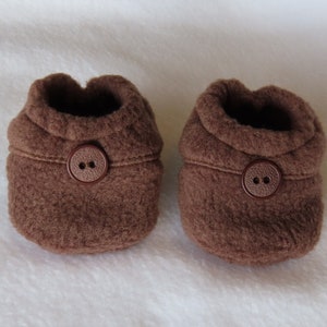 May include: A pair of brown baby booties made from a soft, textured fabric. Each bootie has a rounded shape with a folded cuff and a large, round button fastening. These booties are designed for infants.