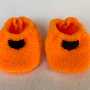 May include: A pair of vibrant orange baby booties, each featuring a black bat-shaped applique. The booties are crafted from a soft, fluffy fabric, ideal for Halloween. The open design allows for easy wear.