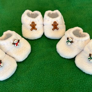 May include: Six pairs of white, fluffy baby booties with a Christmas theme. Each pair has a different decorative button: a gingerbread man or a snowman. The booties are arranged on a green surface.