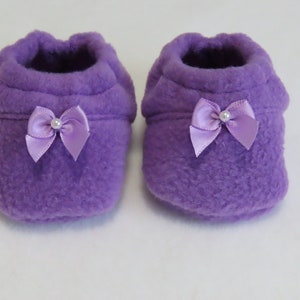May include: A pair of soft, purple baby booties. Each bootie has a satin bow with a pearl accent. The booties are made of a plush material, perfect for keeping little feet warm. A charming and cozy accessory for newborns.