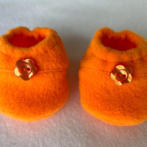 May include: Two bright orange baby booties made from soft fleece. Each bootie has a flower-shaped button in a gold tone. The booties are designed to keep a baby's feet warm and cosy.