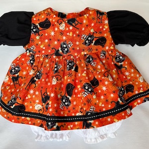 Peut inclure: Une robe avec des manches bouffantes noires courtes et un corsage orange orné d'un motif répétitif de chats noirs, de bonbons maïs et d'étoiles blanches. La jupe présente une bande noire horizontale avec des coutures blanches et des culottes bouffantes blanches sont visibles en dessous.