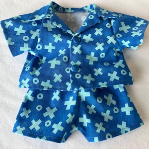 Peut inclure: Un ensemble de pyjama bleu pour poupée, composé d'une chemise boutonnée et d'un short. Le tissu présente un motif de croix et de cercles bleu clair sur un fond bleu plus foncé. La chemise a un col et deux boutons.