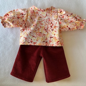 Peut inclure: Une tenue de poupée comprenant un haut à imprimé floral et un pantalon en velours côtelé. Le haut a un fond crème avec des motifs floraux roses, oranges et rouges et des manches bouffantes. Le pantalon est de couleur bordeaux unie.