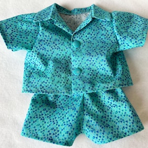 Peut inclure: Un ensemble de poupée turquoise composé d'une chemise à manches courtes boutonnée et d'un short assorti. Le tissu est orné de petits triangles bleus. La chemise a un col et deux boutons turquoise. Le short a un design simple et ajusté.