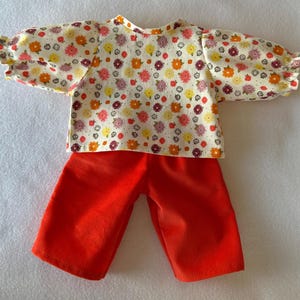 Peut inclure: Un ensemble pour poupée composé d'un haut à imprimé floral et d'un short en velours côtelé orange vif. Le haut présente un fond blanc orné d'un motif de fleurs colorées. Le short est de couleur orange vif unie.