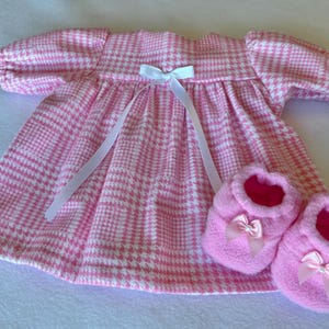 May include: A pink and white checked baby dress with a white bow and matching pink baby booties with bows. The dress has short, puffed sleeves and a ribbon tie. The booties are soft and fluffy.