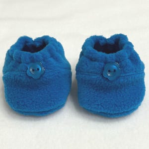 May include: A pair of blue baby booties made from a soft, plush fabric. Each bootie has a flower-shaped button detail. These booties are designed to keep a baby's feet warm and comfortable.