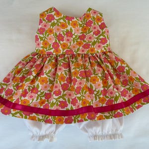 May include: A sleeveless dress featuring a floral pattern of pink, orange, and light pink flowers with green leaves on a white background, accented by a horizontal dark pink ribbon near the hem, paired with white ruffled bloomers underneath.