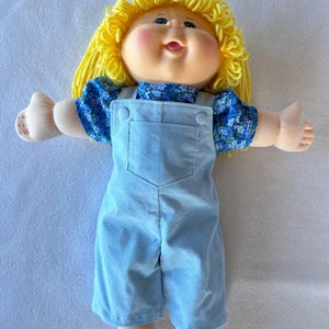 May include: A vintage Cabbage Patch Kid doll with a smiling face, rosy cheeks, and blue eyes. The doll has yellow yarn hair and is wearing a blue floral print shirt under light blue corduroy dungarees. The doll's arms are outstretched.