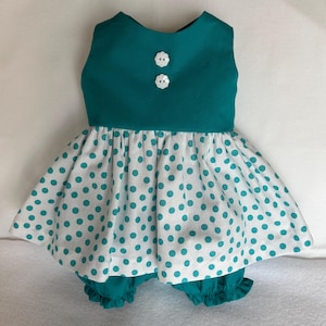 May include: A teal and white polka dot dress with matching bloomers. The sleeveless top has two white flower-shaped buttons. The skirt and bloomers feature a white fabric with teal polka dots. The bloomers have a ruffled hem.