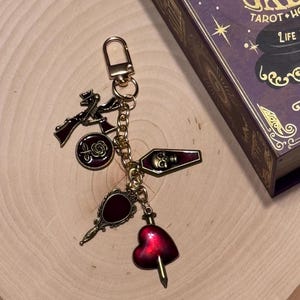 May include: Gold-toned keychain with various charms, including a rose, a coffin with a skull, a heart pierced by a dagger, and a hand mirror. The charms are in shades of red and gold. The keychain is on a wooden surface.