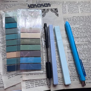 May include: An assortment of stationery items on a printed paper background. The items include a pack of colorful sticky notes, a black pen, two highlighters in light purple and light blue, and a blue pen.