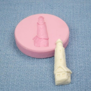 May include: A white plastic lighthouse figurine with a pink silicone mold in the background. The lighthouse has a white base and a tall, narrow tower with a small window.