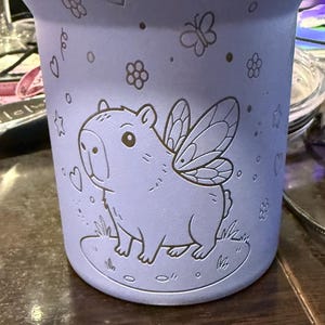 May include: A light purple tumbler with a capybara design, complete with fairy wings, standing on grass. The tumbler is decorated with etched hearts, flowers, butterflies, and stars. The cylindrical tumbler appears to be made of metal.