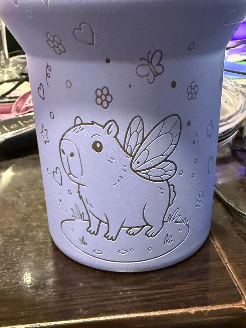 May include: A light purple tumbler with a capybara design, complete with fairy wings, standing on grass. The tumbler is decorated with etched hearts, flowers, butterflies, and stars. The cylindrical tumbler appears to be made of metal.