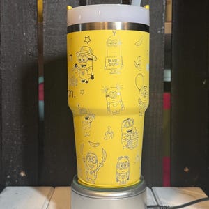 May include: A bright yellow stainless steel tumbler featuring a repeating pattern of black line art illustrations of Minions and bananas. The tumbler has a white lid with a clear straw and text that reads I'M WITH STUPID on one of the characters.