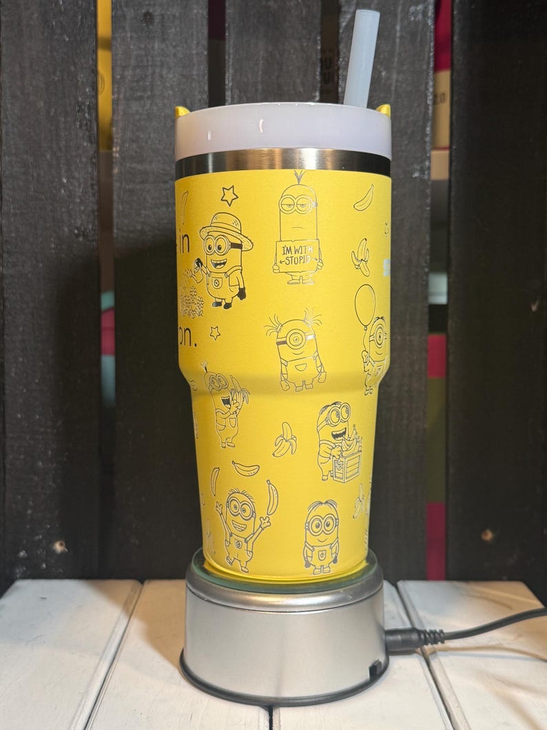 May include: A bright yellow stainless steel tumbler featuring a repeating pattern of black line art illustrations of Minions and bananas. The tumbler has a white lid with a clear straw and text that reads I'M WITH STUPID on one of the characters.