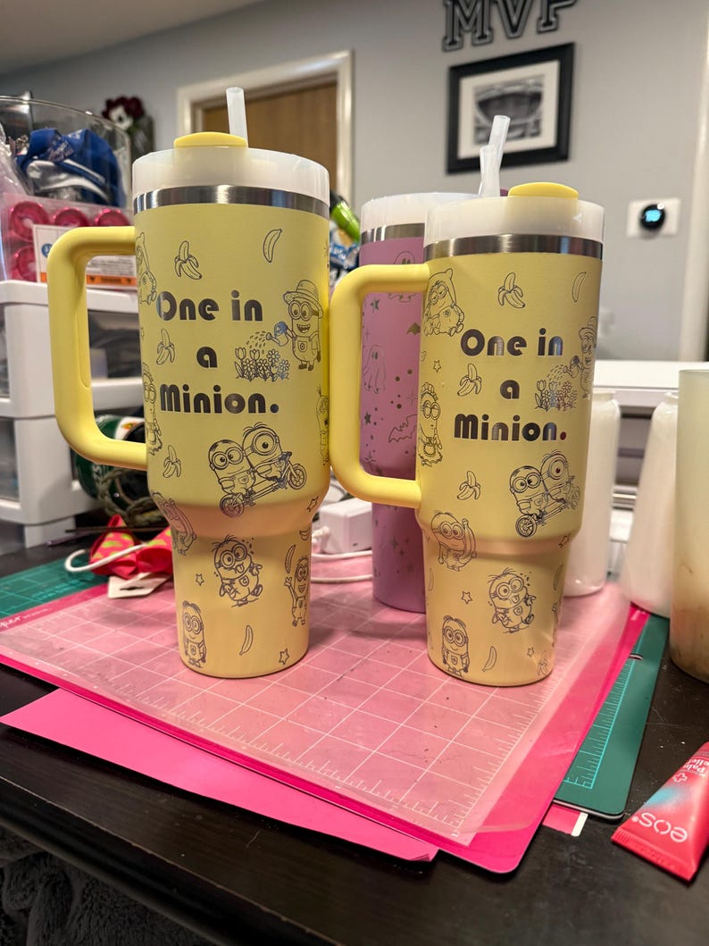 Pu&ograve; includere: Due bicchieri termici gialli con manici e cannucce, caratterizzati da disegni a linee nere di personaggi Minion, banane e il testo One in a Minion. I bicchieri hanno una base affusolata, un coperchio bianco con tappo giallo e una cannuccia inserita.