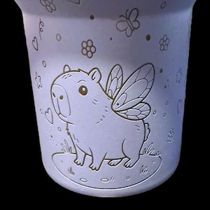 May include: A light purple ceramic mug with a whimsical design. It features a capybara with fairy wings, surrounded by hearts, flowers, and butterflies. The mug has a slightly flared shape.