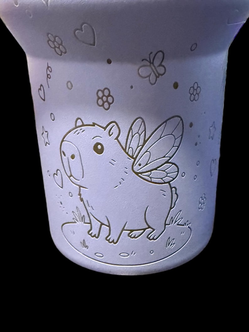 May include: A light purple ceramic mug with a whimsical design. It features a capybara with fairy wings, surrounded by hearts, flowers, and butterflies. The mug has a slightly flared shape.