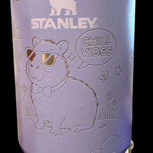 May include: A light purple Stanley travel mug with a handle, featuring an engraved capybara wearing sunglasses and a bow tie. The mug also has the text "CHILL VIBES" and the Stanley logo.