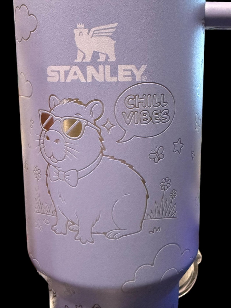 May include: A light purple Stanley travel mug with a handle, featuring an engraved capybara wearing sunglasses and a bow tie. The mug also has the text "CHILL VIBES" and the Stanley logo.