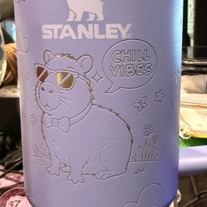 May include: A light purple Stanley mug featuring a capybara wearing sunglasses and a bow tie, with the words "CHILL VIBES" in a speech bubble. The mug also has the Stanley logo and cloud, flower, and star designs.