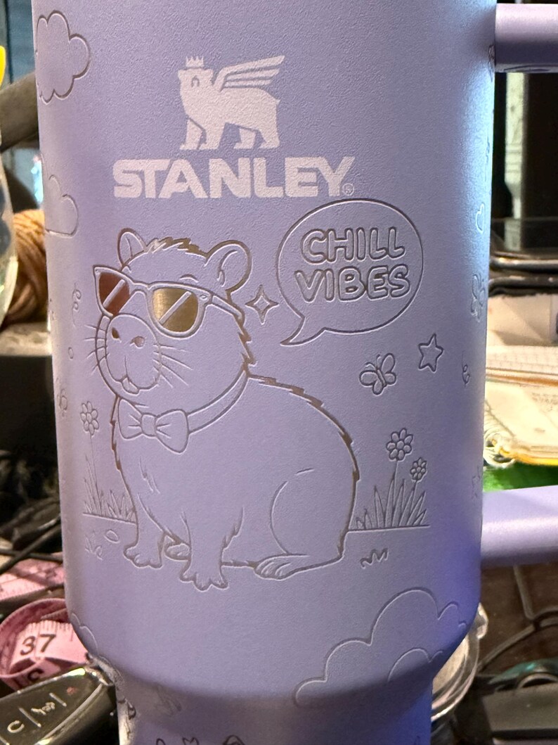 May include: A light purple Stanley mug featuring a capybara wearing sunglasses and a bow tie, with the words "CHILL VIBES" in a speech bubble. The mug also has the Stanley logo and cloud, flower, and star designs.