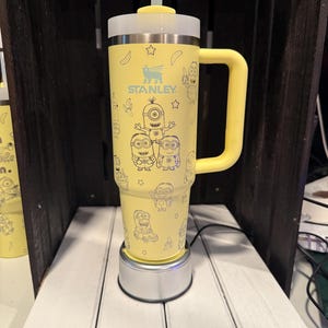 May include: A yellow Stanley tumbler with a handle, a clear straw, and a white lid. The exterior features a repeating blue line-art pattern of Minion characters, bananas, and stars. The Stanley logo is printed in blue on the upper front section.