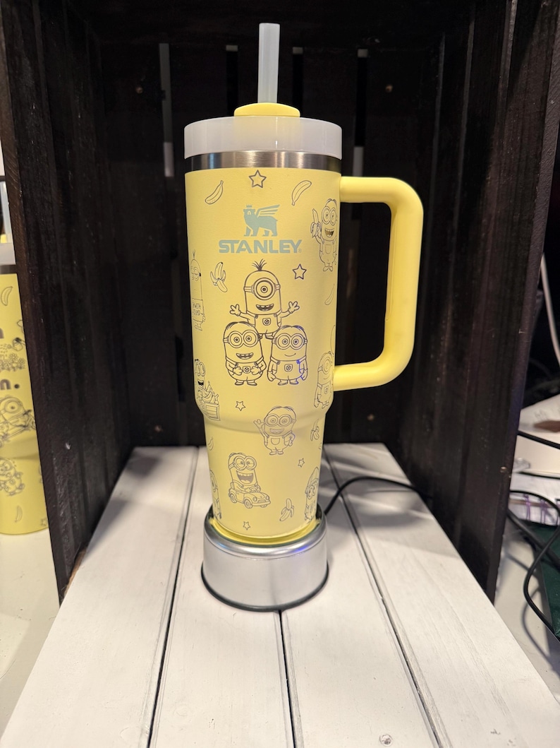 May include: A yellow Stanley tumbler with a handle, a clear straw, and a white lid. The exterior features a repeating blue line-art pattern of Minion characters, bananas, and stars. The Stanley logo is printed in blue on the upper front section.