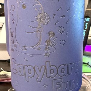 May include: A lilac-coloured tumbler featuring a capybara illustration. The capybara holds a dandelion, with the text "Capybara Fun!" below. The design includes a sun, keys, butterflies, and hearts.