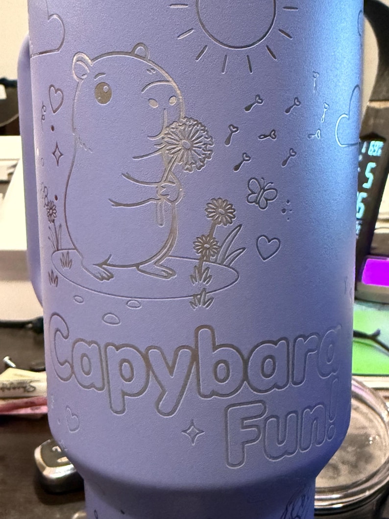 May include: A lilac-coloured tumbler featuring a capybara illustration. The capybara holds a dandelion, with the text "Capybara Fun!" below. The design includes a sun, keys, butterflies, and hearts.