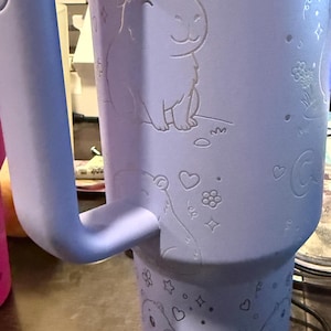 May include: A light purple insulated tumbler with a handle. The tumbler has etched illustrations of capybaras wearing flower crowns, hearts, stars, and flowers. The handle is a matching light purple colour.
