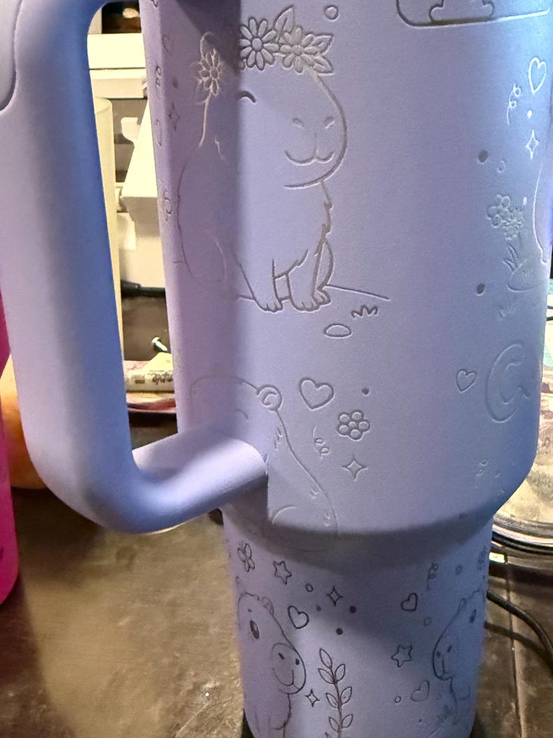 May include: A light purple insulated tumbler with a handle. The tumbler has etched illustrations of capybaras wearing flower crowns, hearts, stars, and flowers. The handle is a matching light purple colour.