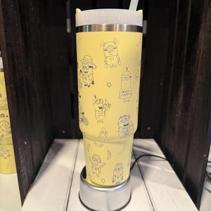 May include: A yellow stainless steel tumbler featuring a repeating pattern of black line art illustrations of Minions and bananas. The tumbler has a white lid with small yellow ears and a clear straw. One Minion holds a sign that reads I'M WITH STUPID.
