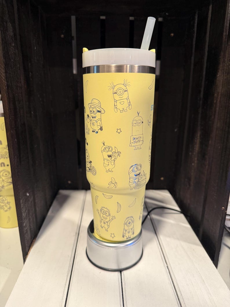 May include: A yellow stainless steel tumbler featuring a repeating pattern of black line art illustrations of Minions and bananas. The tumbler has a white lid with small yellow ears and a clear straw. One Minion holds a sign that reads I'M WITH STUPID.