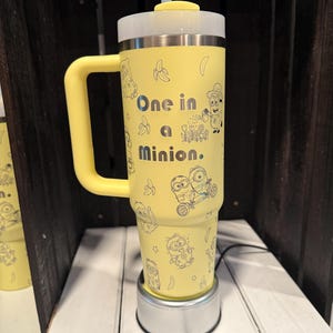 May include: A yellow insulated tumbler with a handle, a clear lid, and a straw. The exterior features black line art of Minion characters, bananas, and flowers, with the text "One in a Minion." printed in the center in a bold, dark font.