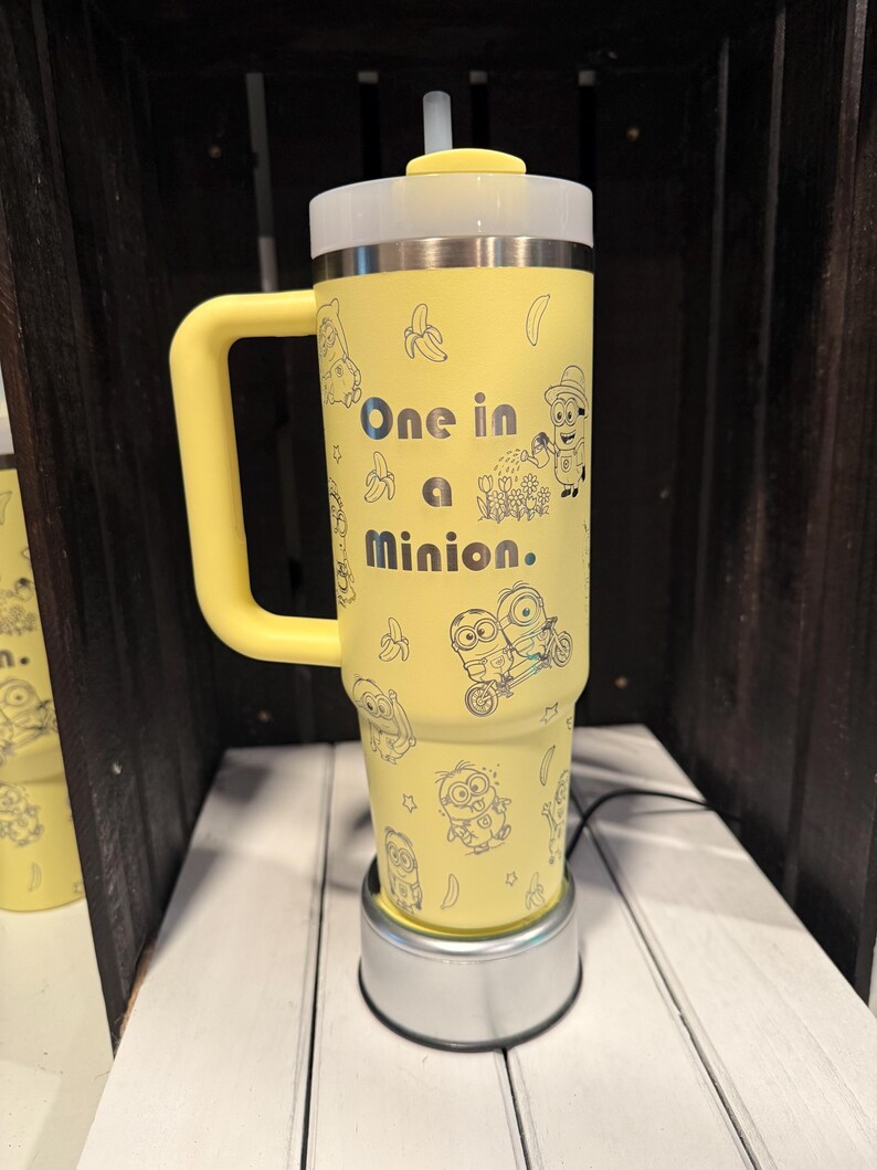 May include: A yellow insulated tumbler with a handle, a clear lid, and a straw. The exterior features black line art of Minion characters, bananas, and flowers, with the text "One in a Minion." printed in the center in a bold, dark font.