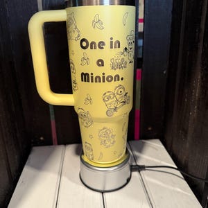 K&ouml;nnte beinhalten: Ein gelber Isolierbecher mit Henkel, verziert mit schwarzen Strichzeichnungen von Minion-Figuren und Bananen. Der Text "One in a Minion." ist in Schwarz auf der Vorderseite aufgedruckt. Der Becher hat einen hellgelben Deckel mit Strohhalm und steht auf einem silbernen Sockel.