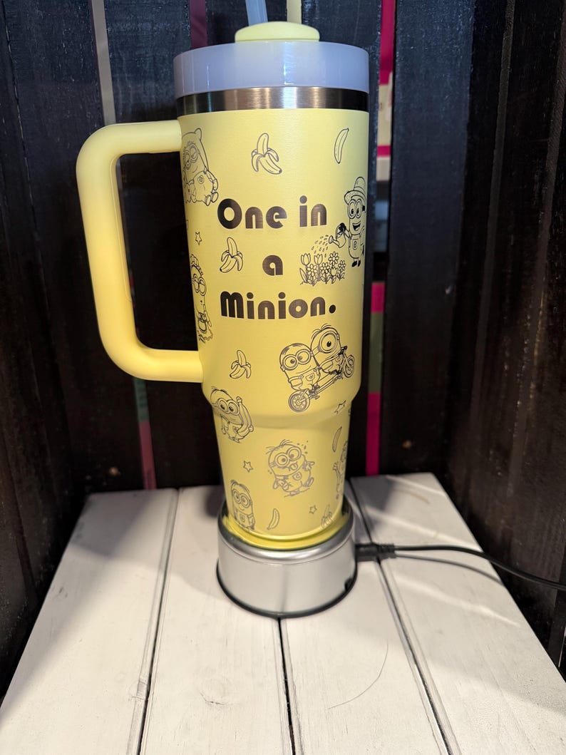 K&ouml;nnte beinhalten: Ein gelber Isolierbecher mit Henkel, verziert mit schwarzen Strichzeichnungen von Minion-Figuren und Bananen. Der Text "One in a Minion." ist in Schwarz auf der Vorderseite aufgedruckt. Der Becher hat einen hellgelben Deckel mit Strohhalm und steht auf einem silbernen Sockel.