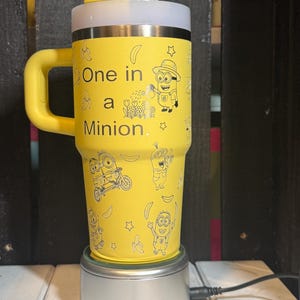 May include: A yellow tumbler with a handle, a white lid, and a clear straw. The tumbler features black line art of Minions, bananas, and stars, with the text "One in a Minion." printed on the front. The cup is resting on a circular silver base.