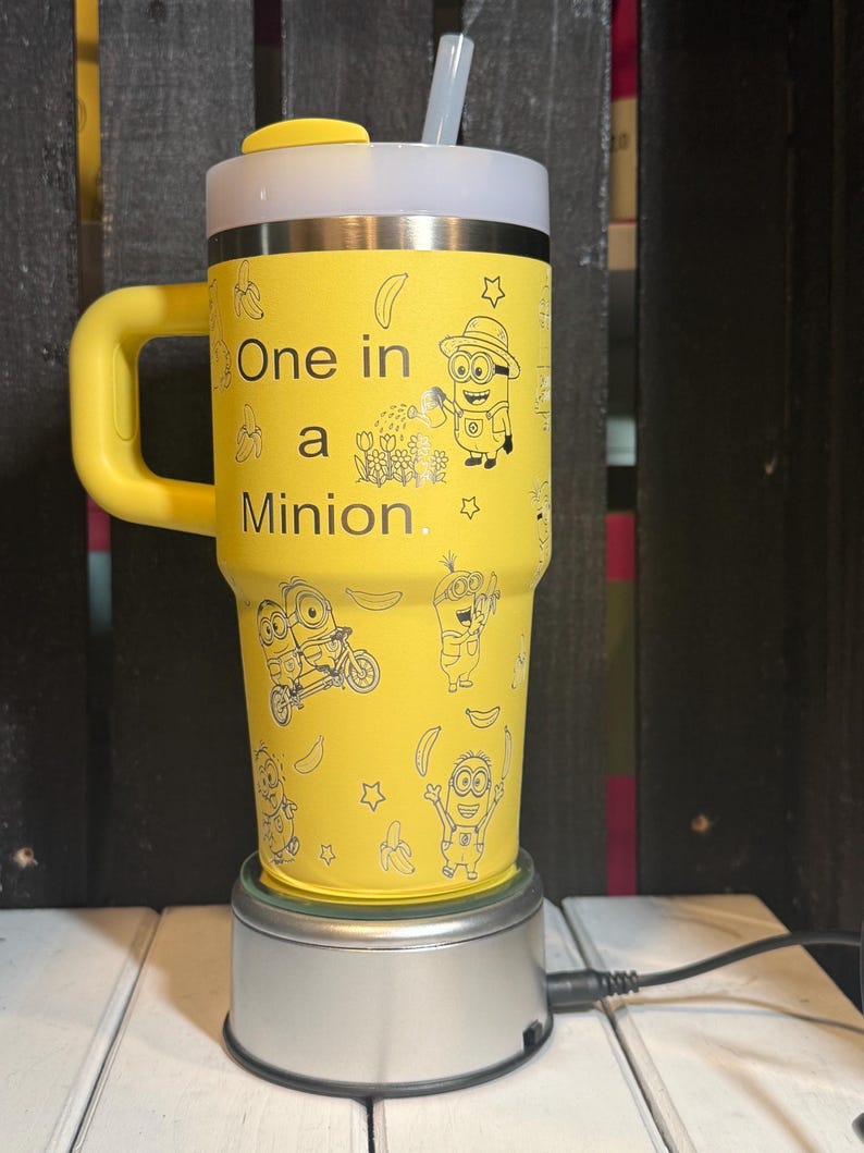 May include: A yellow tumbler with a handle, a white lid, and a clear straw. The tumbler features black line art of Minions, bananas, and stars, with the text "One in a Minion." printed on the front. The cup is resting on a circular silver base.
