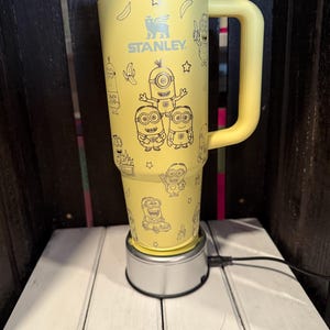 K&ouml;nnte beinhalten: Ein gelber Stanley-Becher mit einem schwarzen Linienmuster aus Minions, Bananen und Sternen. Der Isolierbecher verf&uuml;gt &uuml;ber einen gro&szlig;en seitlichen Griff, einen metallischen Rand und einen hellen Deckel. Der Markenname STANLEY ist in Schwarz aufgedruckt.