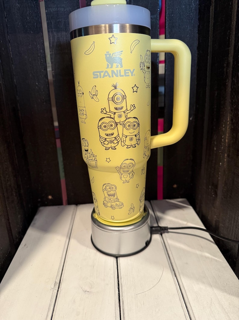 K&ouml;nnte beinhalten: Ein gelber Stanley-Becher mit einem schwarzen Linienmuster aus Minions, Bananen und Sternen. Der Isolierbecher verf&uuml;gt &uuml;ber einen gro&szlig;en seitlichen Griff, einen metallischen Rand und einen hellen Deckel. Der Markenname STANLEY ist in Schwarz aufgedruckt.