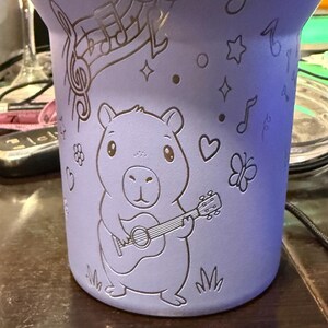 May include: A lilac-coloured tumbler with etched designs. The tumbler features a capybara playing a guitar, musical notes, stars, hearts, and a butterfly. The design is etched into the surface of the tumbler.