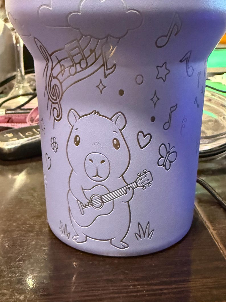 May include: A lilac-coloured tumbler with etched designs. The tumbler features a capybara playing a guitar, musical notes, stars, hearts, and a butterfly. The design is etched into the surface of the tumbler.