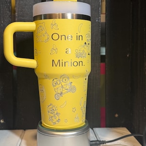 May include: A bright yellow insulated tumbler with a handle, a clear lid, and a straw. The exterior features black line art of Minion characters, bananas, and flowers, with the text One in a Minion printed in the center of the cup.