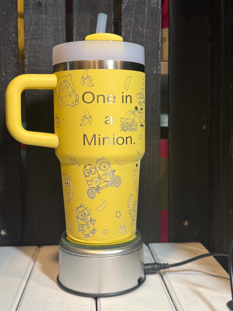 May include: A bright yellow insulated tumbler with a handle, a clear lid, and a straw. The exterior features black line art of Minion characters, bananas, and flowers, with the text One in a Minion printed in the center of the cup.