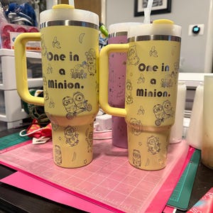 May include: Two yellow insulated tumblers with handles and straws, featuring black line art of Minion characters, bananas, and the text One in a Minion. The tumblers have a tapered base, a white lid with a yellow stopper, and a clear straw inserted.